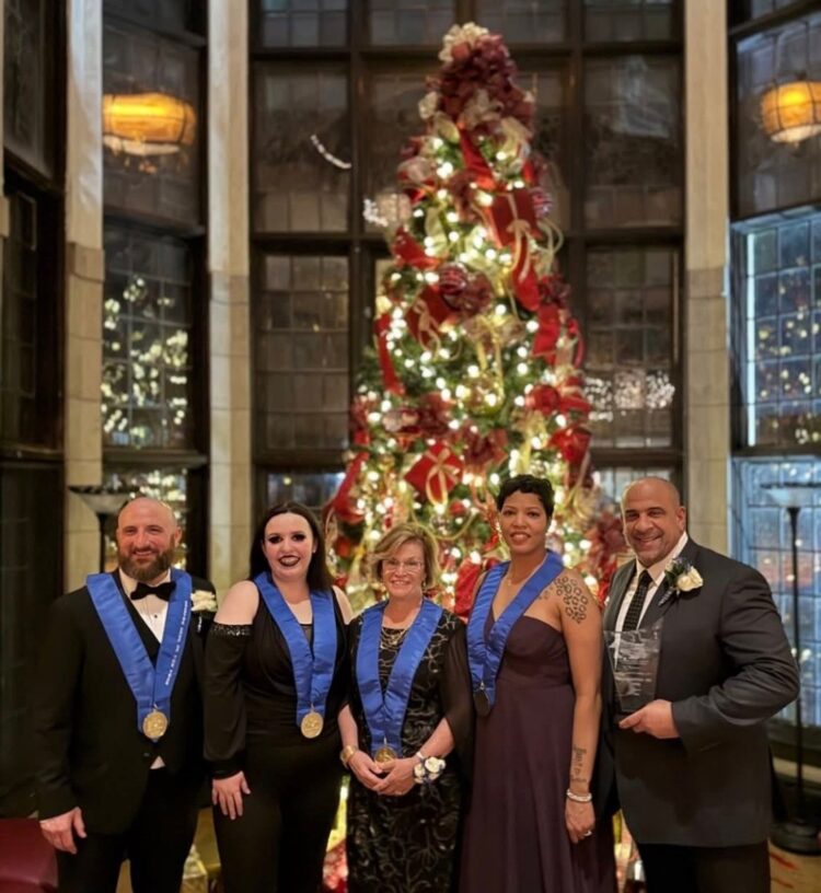 Five formally dressed people with blue ribbon medals stand smiling in front of a decorated, lit Christmas tree in an elegant room with large windows. One person on the right holds a glass award.