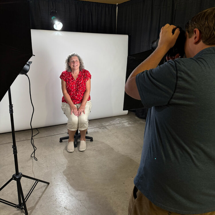 A woman in a red floral shirt sits on a stool, smiling, in front of a white backdrop while a photographer takes her picture in a studio with professional lighting.