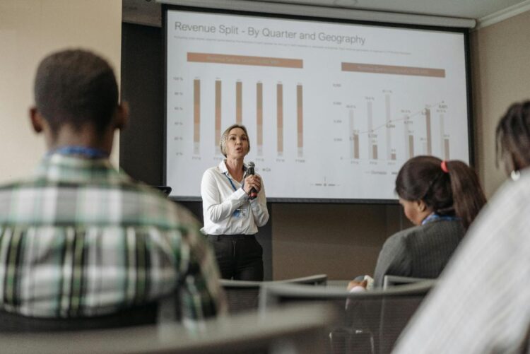 A woman stands and speaks in front of an audience, presenting a slide with charts titled Revenue Split – By Quarter and Geography in a conference room. Several people listen attentively.