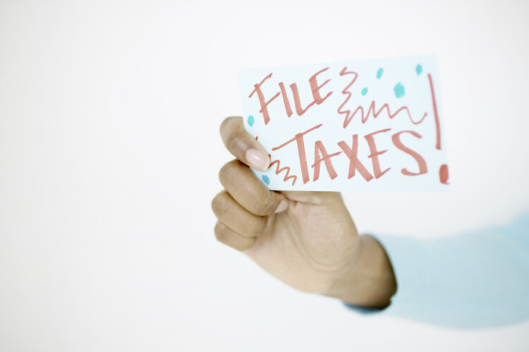 A hand holding a card with the words File Taxes! written in bold, colorful letters, with decorative squiggles and dots, against a plain white background.