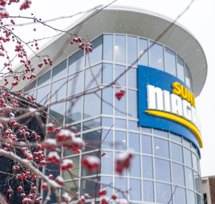 Snow-covered red berries on branches in the foreground, with a modern glass building and a partially visible SUNY Niagara sign in the background, on a snowy day.