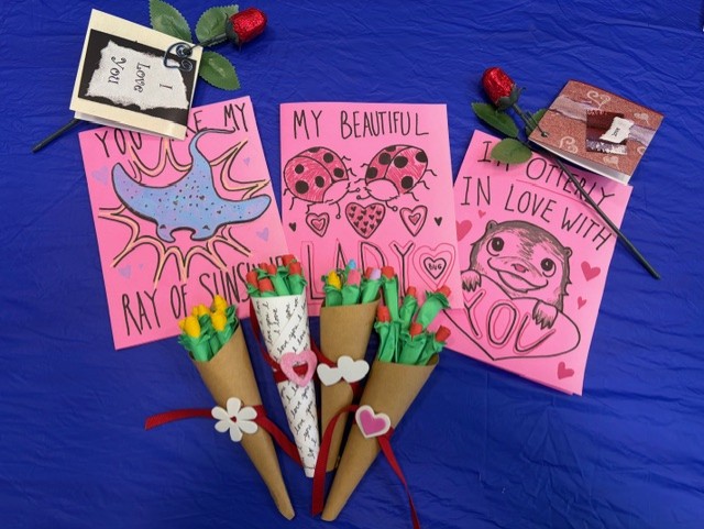 Three handmade pink Valentine cards with cute animal drawings and messages are displayed on a blue surface, surrounded by paper roses and three small paper bouquets with rolled paper flowers and hearts.
