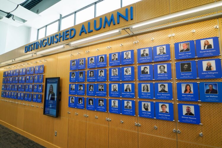 A wall display titled “Distinguished Alumni” features rows of blue plaques with black and white portraits, names, and details of notable graduates mounted on a perforated wooden wall.