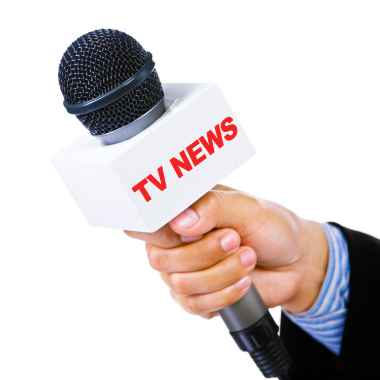 A person’s hand holding a microphone with a white square labeled “TV NEWS” in red letters, ready for an interview or news report, against a white background.