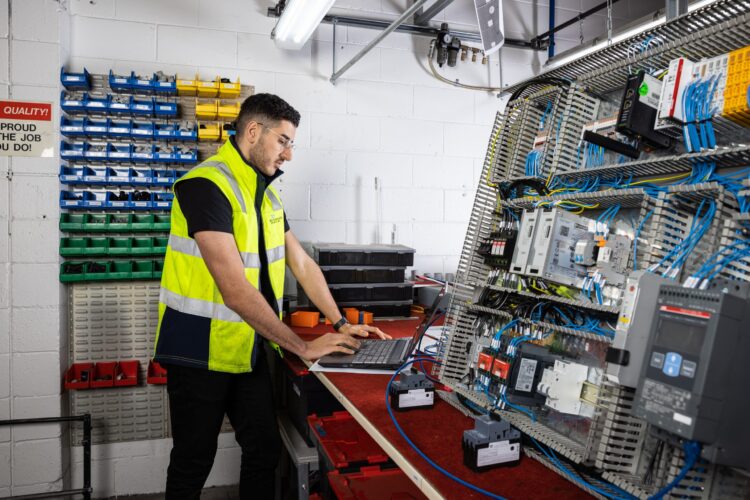 A man wearing a yellow safety vest uses a laptop at a workstation with various electrical components and wiring inside an industrial setting. Colored bins and tools are organized on the wall behind him.