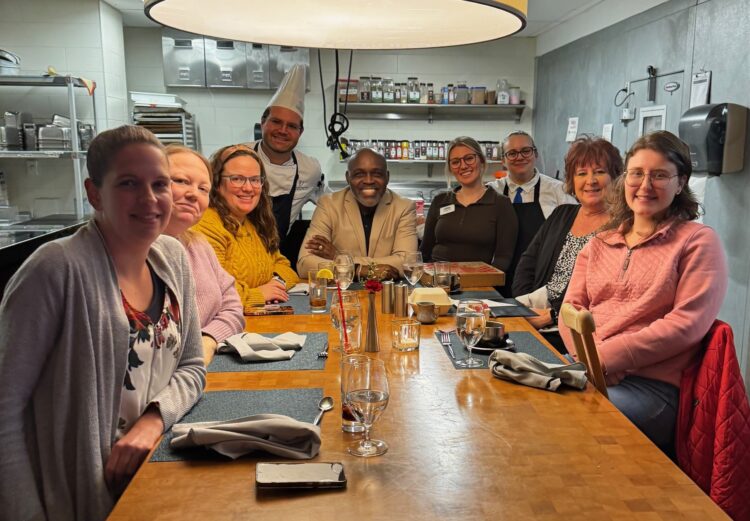 A group of people sit smiling around a restaurant table, with food and drinks in front of them. Two chefs in white uniforms stand at the back. The setting appears warm and friendly in a modern kitchen.