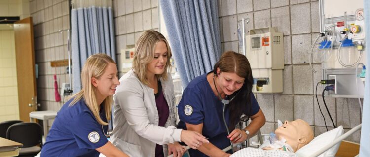 Two nursing students and an instructor practice medical procedures on a hospital simulation mannequin, smiling and engaged in a clinical skills classroom setting.
