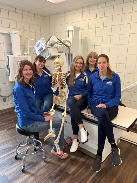 Five women in matching blue shirts sit and stand around a human skeleton model in a medical or radiology room with white tiles and equipment in the background. They are all smiling at the camera.
