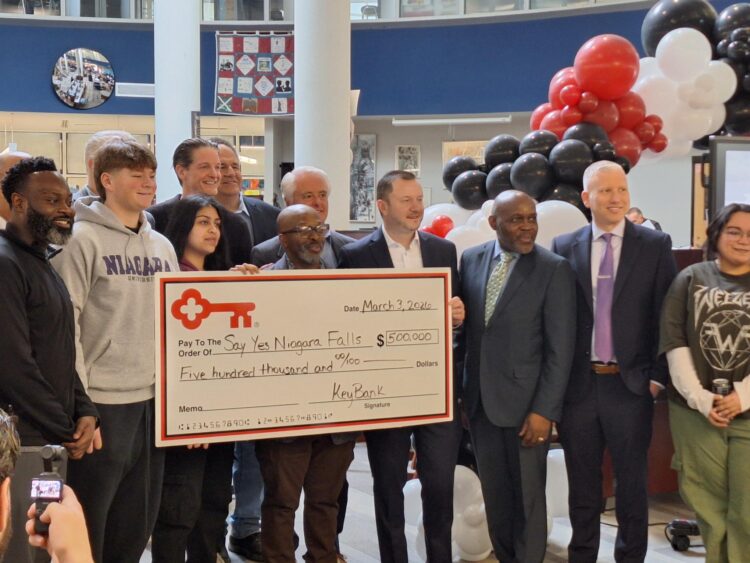 A group of people stands together, smiling and holding an oversized check for $500,000 made out to Say Yes Niagara Falls from KeyBank. Red, white, and black balloons decorate the background.