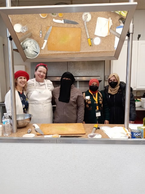 Five women stand behind a kitchen counter with baking supplies, smiling for a group photo. Above them is a mirror reflecting the countertop, utensils, and ingredients.