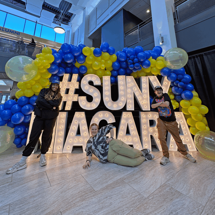 Three people pose next to a large lit-up #SUNY NIAGARA sign, surrounded by blue, yellow, and clear balloons, inside a modern building with a high ceiling and large windows.