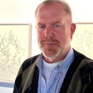 A middle-aged man with a gray beard and short hair wears a checked shirt and a dark sweater, standing indoors in front of framed minimalist line art drawings.