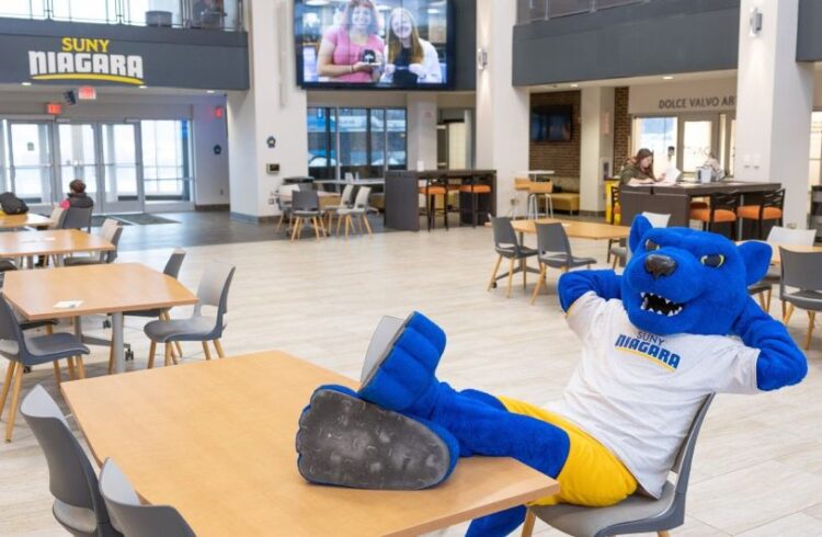 A blue panther mascot in a SUNY Niagara shirt and yellow shorts relaxes with feet up on a table in a spacious, modern campus dining area with empty tables and chairs. A large screen shows people on a video call.