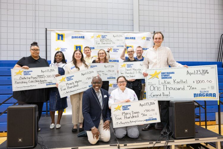 A group of people smile and pose on a stage, holding large ceremonial checks for various dollar amounts. A backdrop displays SUNY Niagara and other logos, indicating a student showcase or award event.