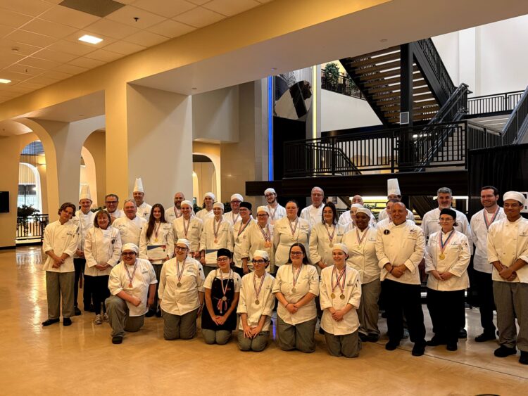 A large group of chefs and culinary students, dressed in white uniforms and chef hats, pose together indoors in a spacious lobby area with a staircase and arches in the background.