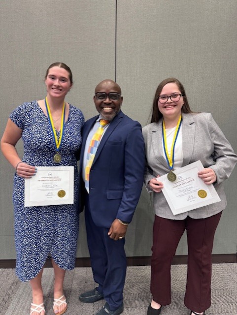 Three people stand indoors smiling at the camera. Two women, each wearing a medal and holding a certificate, stand on either side of a man in a blue suit and colorful tie. The background is a gray wall.