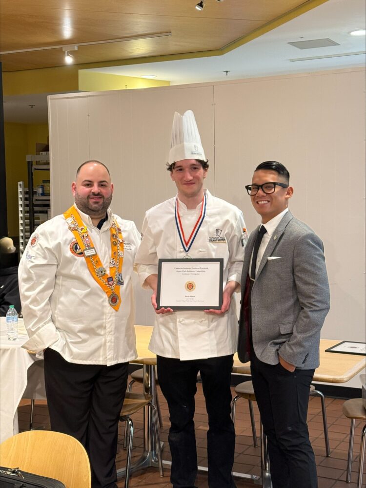 Three men pose indoors. The man in the middle wears a chef’s uniform, chef’s hat, a medal, and holds a framed certificate. The man on the left wears a white coat with a decorative sash. The man on the right wears a gray suit.