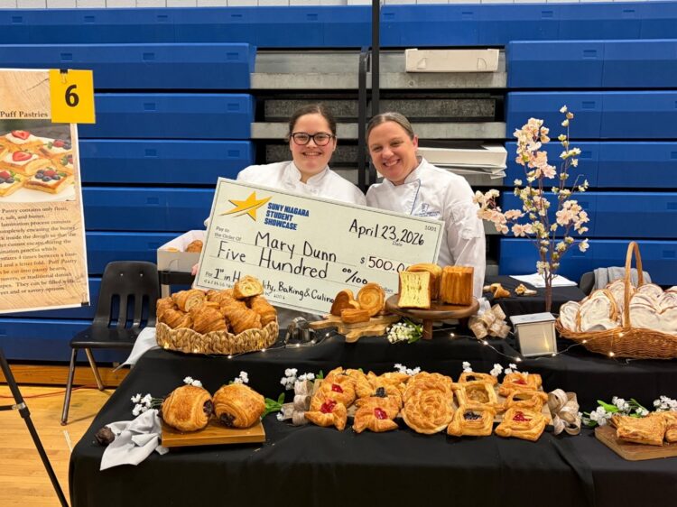 Two women in chef uniforms smile while holding a large $500 check at a table filled with assorted pastries and breads, with blue bleachers and a yellow sign in the background.