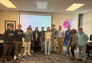 A group of twelve people stand side by side, smiling, in a classroom with computers, a chart on the wall, and a projector screen displaying content in the background.