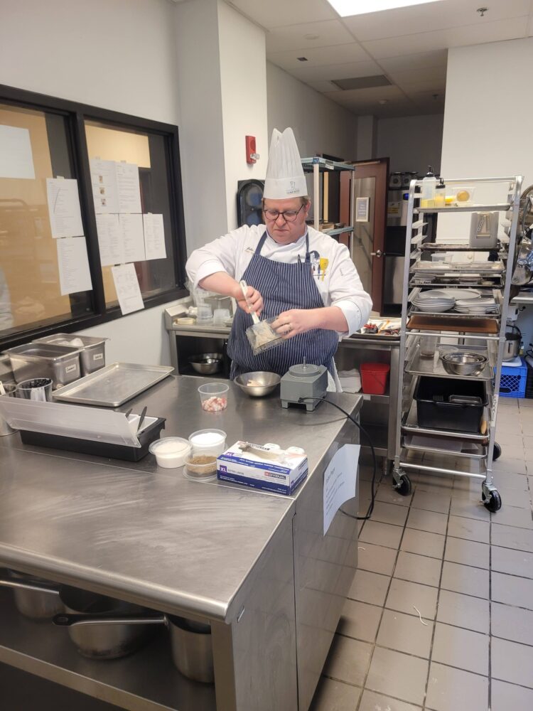A chef in a white uniform and tall hat mixes ingredients in a bowl at a stainless steel kitchen workstation, surrounded by various bowls, utensils, and cooking equipment.