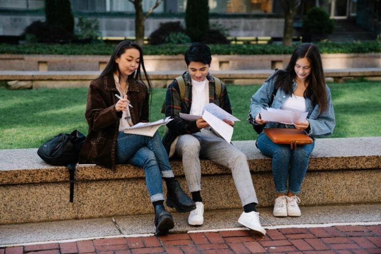 Three students sit on a stone bench outdoors, reading and writing on papers. They appear focused on their work, with backpacks and bags beside them, and a green lawn in the background.