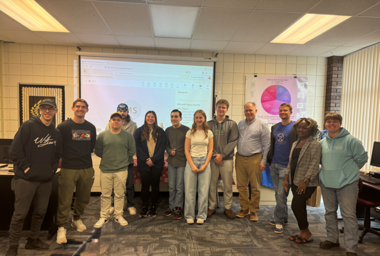A group of 12 people stand side by side, smiling at the camera in a classroom. Behind them is a projector screen displaying a website, along with a colorful poster and computer equipment.