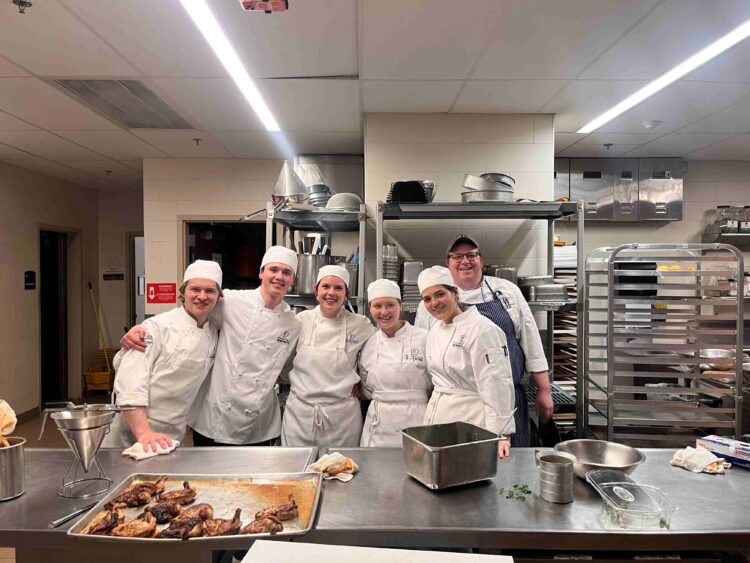 Six people in white chef uniforms and hats stand together smiling in a commercial kitchen, surrounded by metal equipment, trays, and prepared roasted chickens on a counter in the foreground.