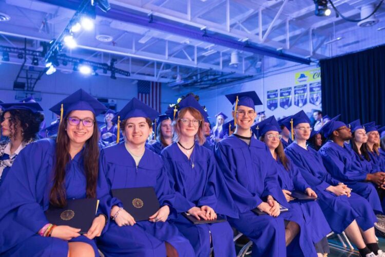 A group of graduates in blue caps and gowns sit in rows, smiling and holding diplomas during a graduation ceremony inside a gymnasium. Banners hang on the wall in the background.