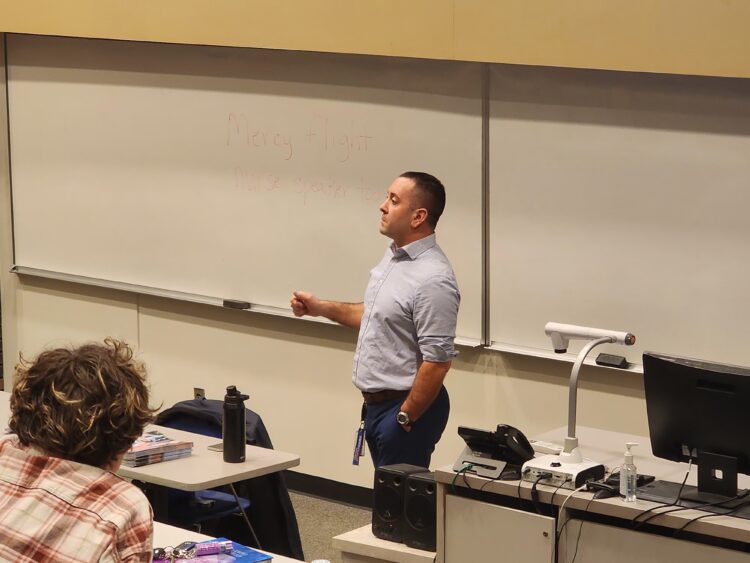 A man in a light blue shirt stands and speaks in front of a classroom, next to a whiteboard with faint writing. Students sit at desks with notebooks and water bottles in the foreground.