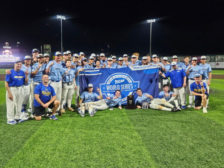 A large group of baseball players and coaches in uniforms pose together on a field at night, smiling and holding up a banner that reads NJCAA Division II World Series Champion. Trophies are displayed in front.