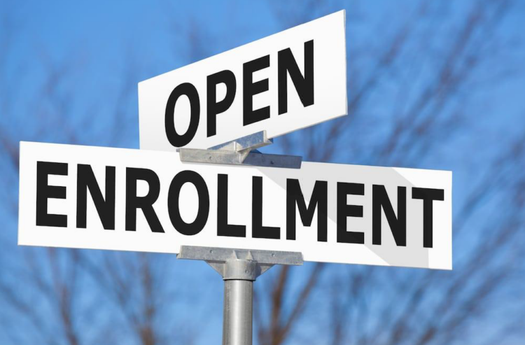 A close-up of two street-style signs reading OPEN and ENROLLMENT against a blue sky with blurred tree branches in the background.
