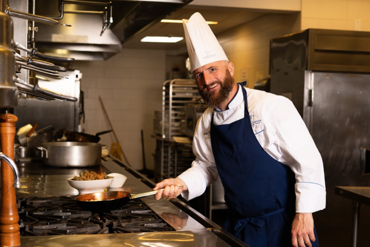 A smiling chef in a white hat and blue apron cooks with a copper pan on a stove in a professional kitchen, with pots, bowls, and cooking equipment visible in the background.