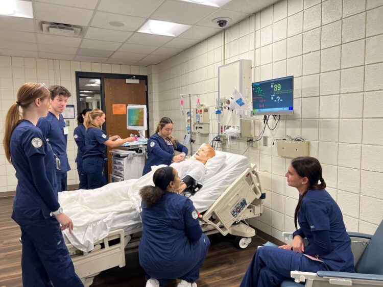 Six nursing students in blue scrubs practice medical procedures on a mannequin in a hospital bed, surrounded by medical equipment and monitors, in a clinical classroom setting.