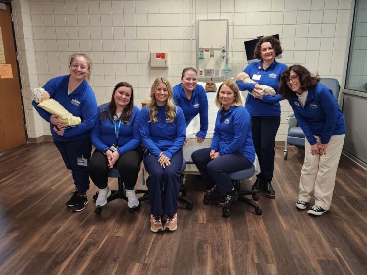Seven women in matching blue shirts pose in a medical room. Two are holding baby mannequins, suggesting a healthcare or training setting. All are smiling and sitting or standing on a wood floor.