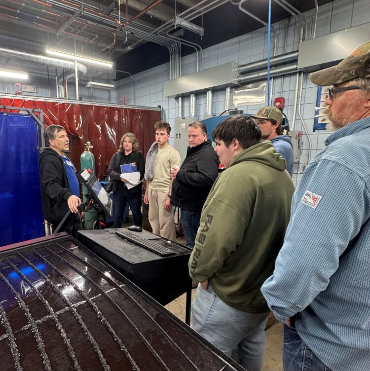 A group of people listens to an instructor explaining equipment in an industrial workshop, surrounded by metal surfaces, pipes, and safety gear. The atmosphere appears educational and hands-on.