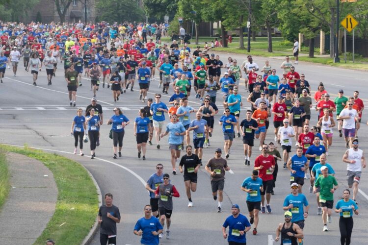 A large crowd of runners in colorful shirts participates in a road race on a wide city street lined with trees, with spectators visible in the background.