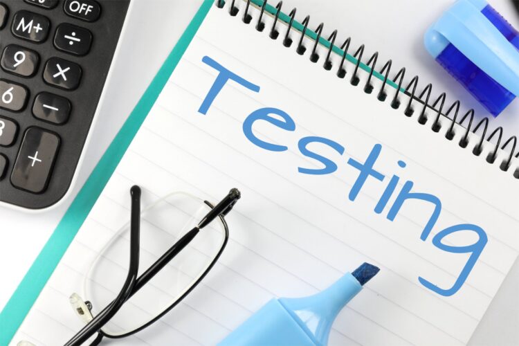 A spiral notebook with the word Testing written in large blue letters. Nearby are a calculator, eyeglasses, a blue highlighter with its cap off, and a blue-and-white eraser.