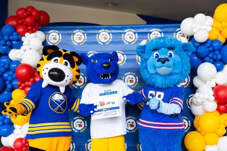 Three colorful sports mascots pose together in front of a balloon arch and branded backdrop. The middle mascot holds a sign. All mascots are smiling and wearing blue-themed sports jerseys.