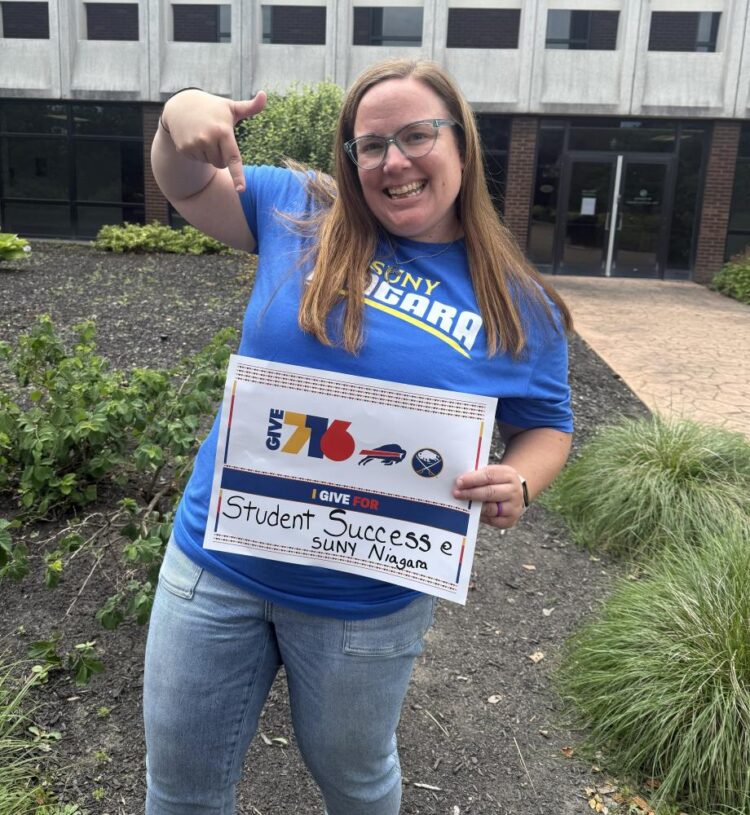 A smiling woman wearing a blue SUNY Niagara shirt stands outside, pointing to a sign that reads, I GIVE FOR Student Success, SUNY Niagara, supporting Give716 and displaying the Buffalo Bills and Sabres logos.
