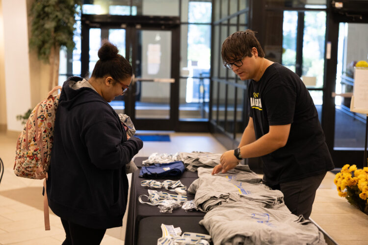 Two people stand at a table with neatly stacked gray t-shirts and lanyards. One person picks up a shirt while the other helps. They are indoors near large glass doors.