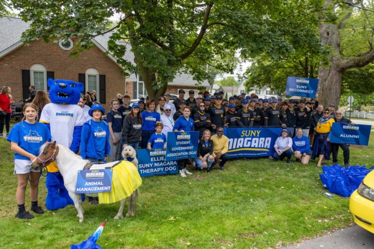 A large group of people, some in Niagara shirts, pose outdoors with a blue bear mascot, therapy dogs, and a small pony. They hold blue SUNY Niagara signs on a lawn with trees and a brick building behind them.