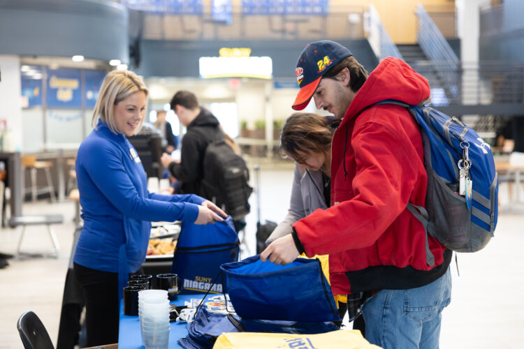 A woman hands a drawstring bag to a man in a red jacket and cap at a table indoors, while others look on. The table displays items and is set in a bright, modern building.
