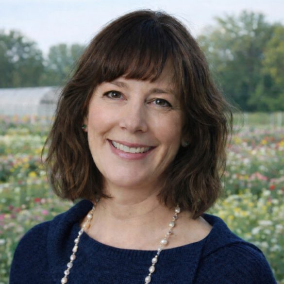 A woman with shoulder-length brown hair and bangs smiles outdoors, wearing a navy sweater and a pearl necklace. A field of flowers and trees are visible in the background under a cloudy sky.