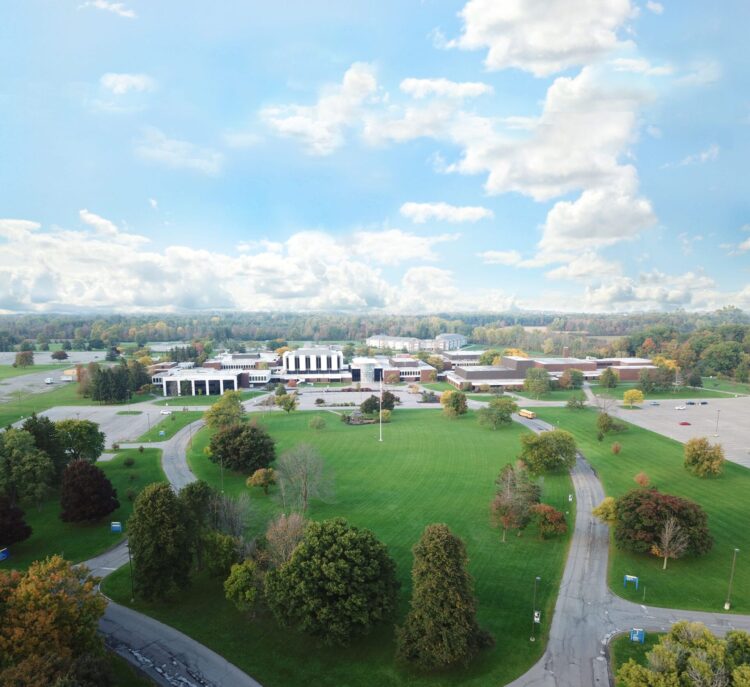 Aerial view of a large school campus surrounded by green lawns, trees with fall foliage, winding roads, and a partly cloudy sky.