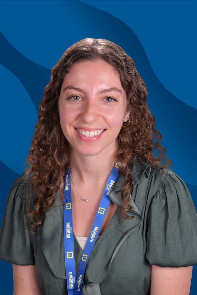 A woman with curly brown hair smiles at the camera. She wears a green blouse and a blue lanyard with yellow text. The background is blue with wavy patterns.