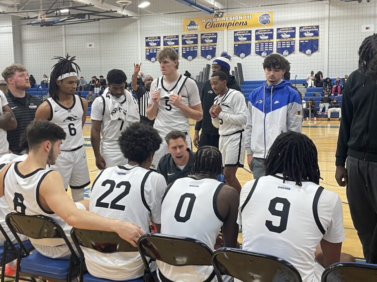 A basketball coach kneels while talking to his seated players during a timeout. Players wear white jerseys with numbers, listening intently. Banners hang on the gym wall in the background.