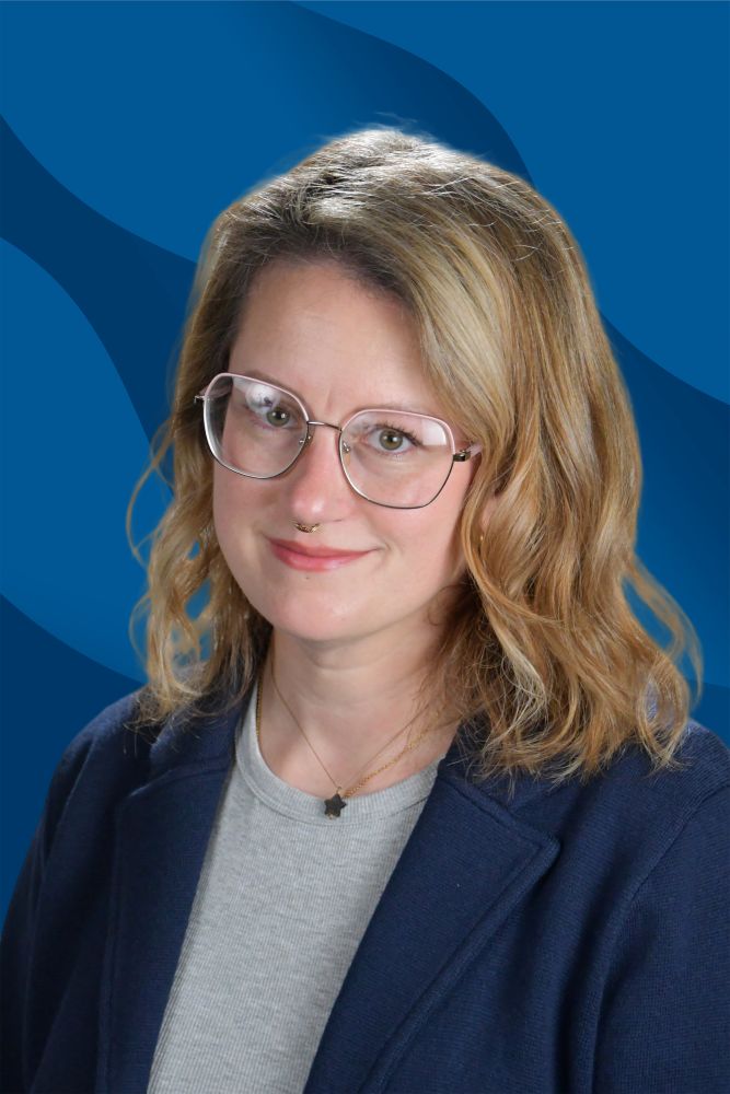 A person with wavy blonde hair, glasses, and a grey shirt under a navy blazer, smiling in front of a blue background with wavy patterns.
