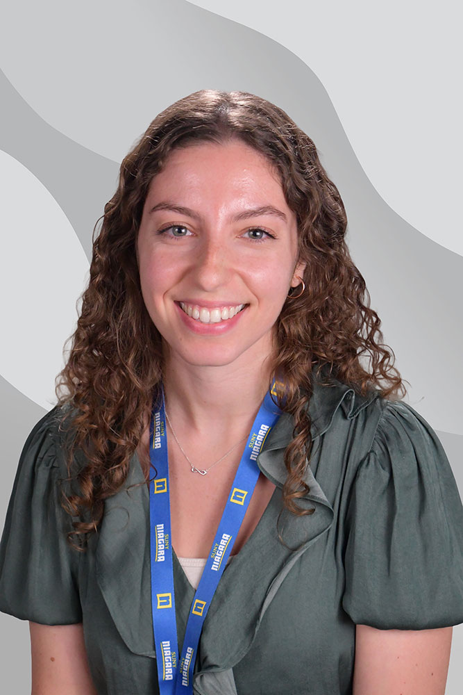 A woman with curly brown hair, wearing a green blouse and a blue lanyard, smiles at the camera. The background has abstract gray and white wavy patterns.