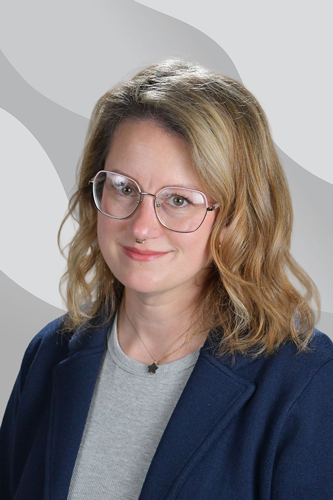 A person with wavy blond hair, large glasses, and a blue blazer over a gray top smiles slightly in front of a light, abstract background.