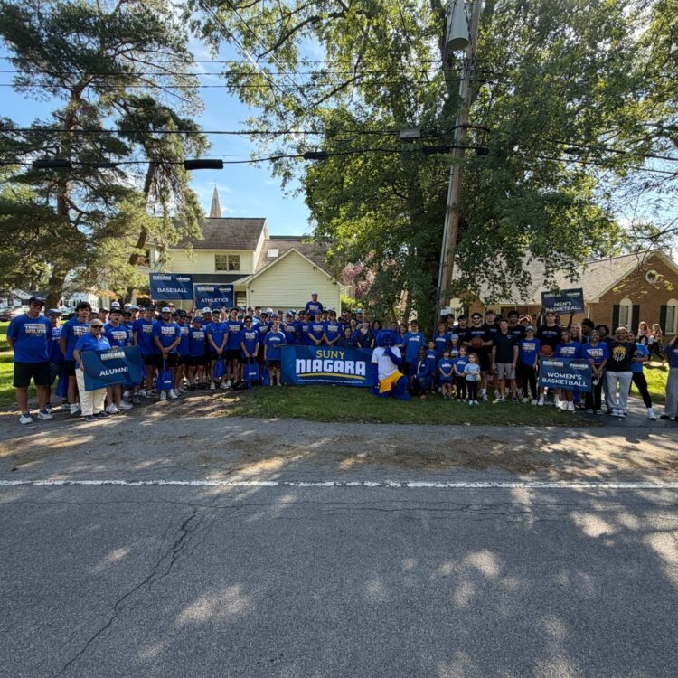 A large group of people in matching blue SUNY Niagara shirts stand outside on a sunny day, holding banners and signs, gathered near trees and houses on a residential street.
